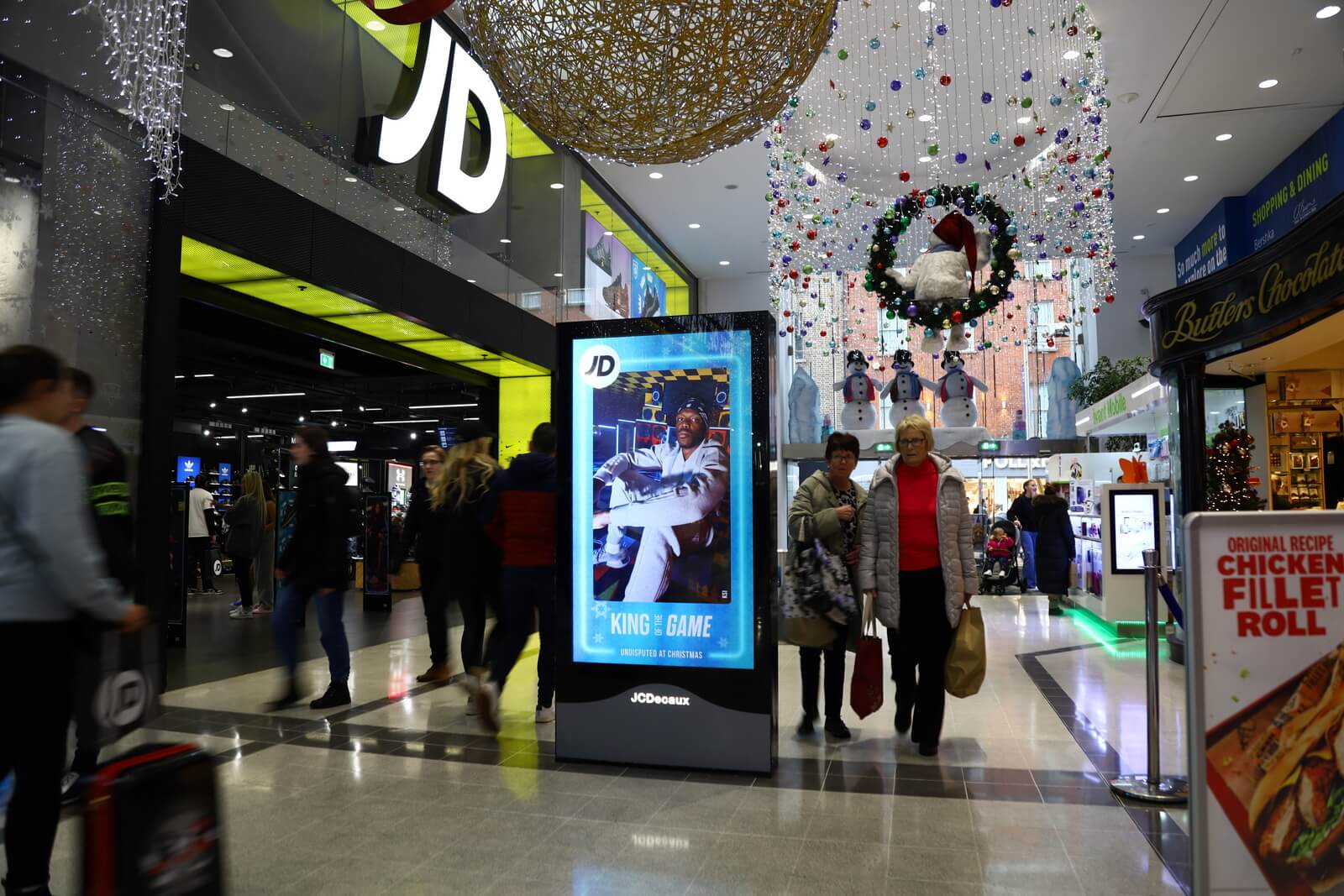 Digital advertising screen outside a JD Sports store in a mall, showcasing festive JD Sports products