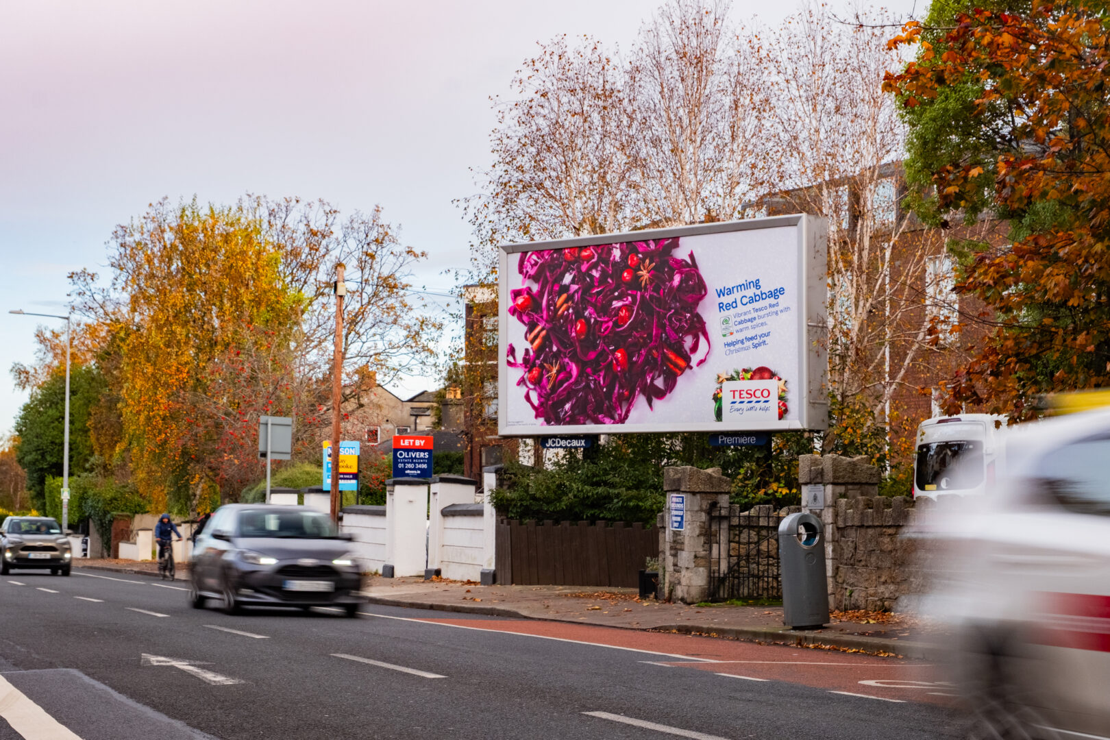 Large roadside billboard featuring a Tesco Christmas campaign with an image of warming red cabbage and festive branding. The billboard is positioned on a busy street with cars passing by, autumn trees in the background, and residential buildings nearby.